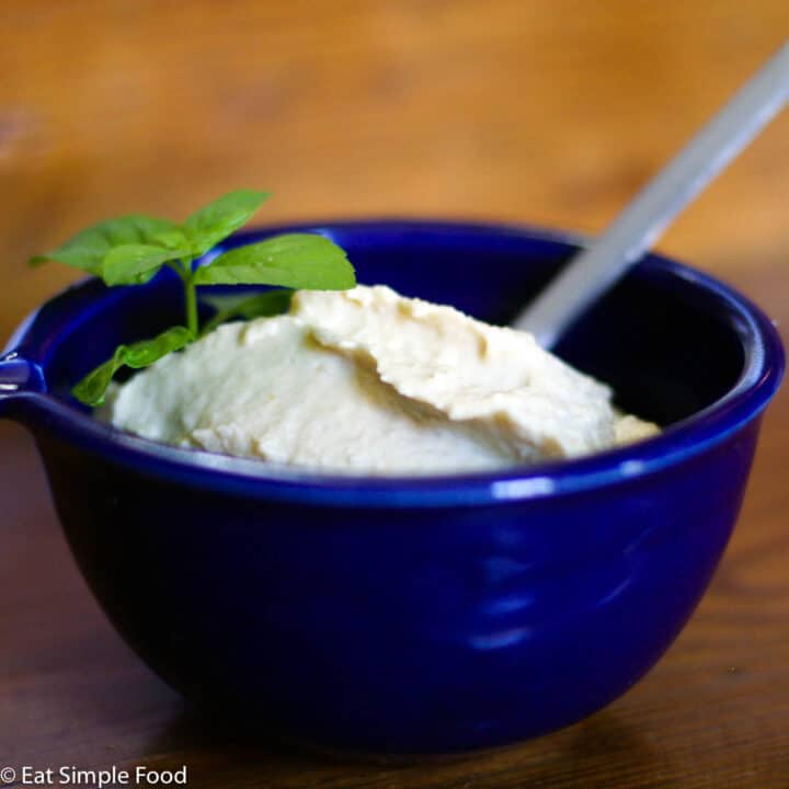 Small blue bowl of off white hummus with a mint garnish. Spoon inserted into hummus.