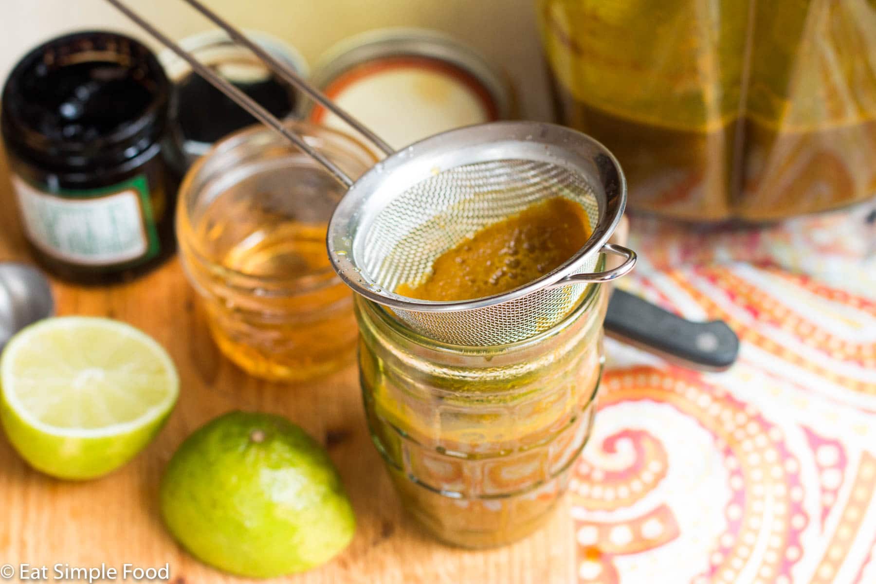 Small glass of brown yellow jamu drink with a small strainer on top for the turmeric and ginger. Lime on side. Small jar of honey on side.