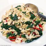White bowl of orzo salad with diced artichoke hearts, red onions, red bell peppers with willted spinach. Silver spoon in the bowl.