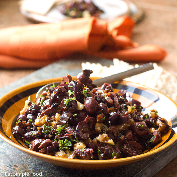Tan bowl of sliced black olives with walnuts and chopped figs mounded in bowl with a spoon and garnished with parsley. side view.