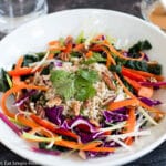 Chopped Kale & Red Cabbage w/ slices of raw red peppers, shaved carrot, sliced green onions w/ brown rice & cilantro in a white bowl with a side of brown peanut salad dressing