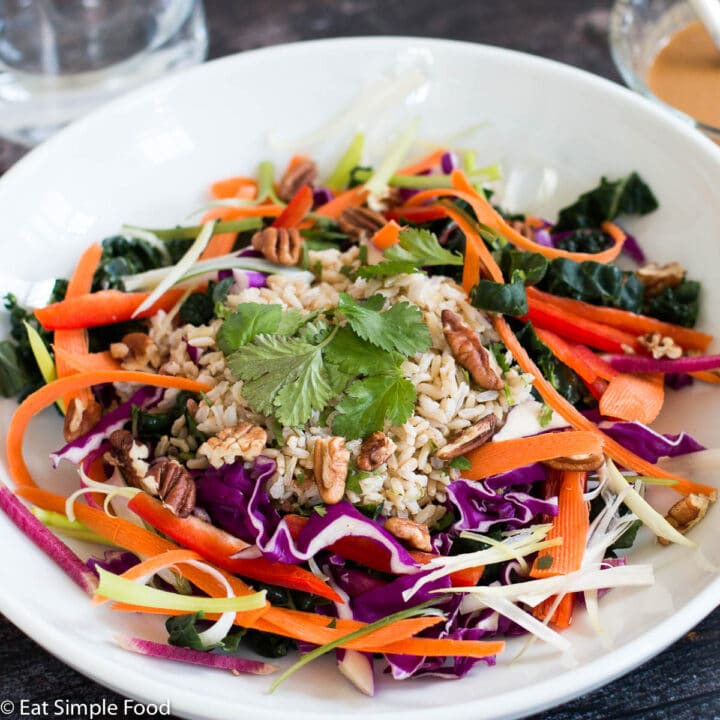 Chopped Kale & Red Cabbage w/ slices of raw red peppers, shaved carrot, sliced green onions w/ brown rice & cilantro in a white bowl with a side of brown peanut salad dressing