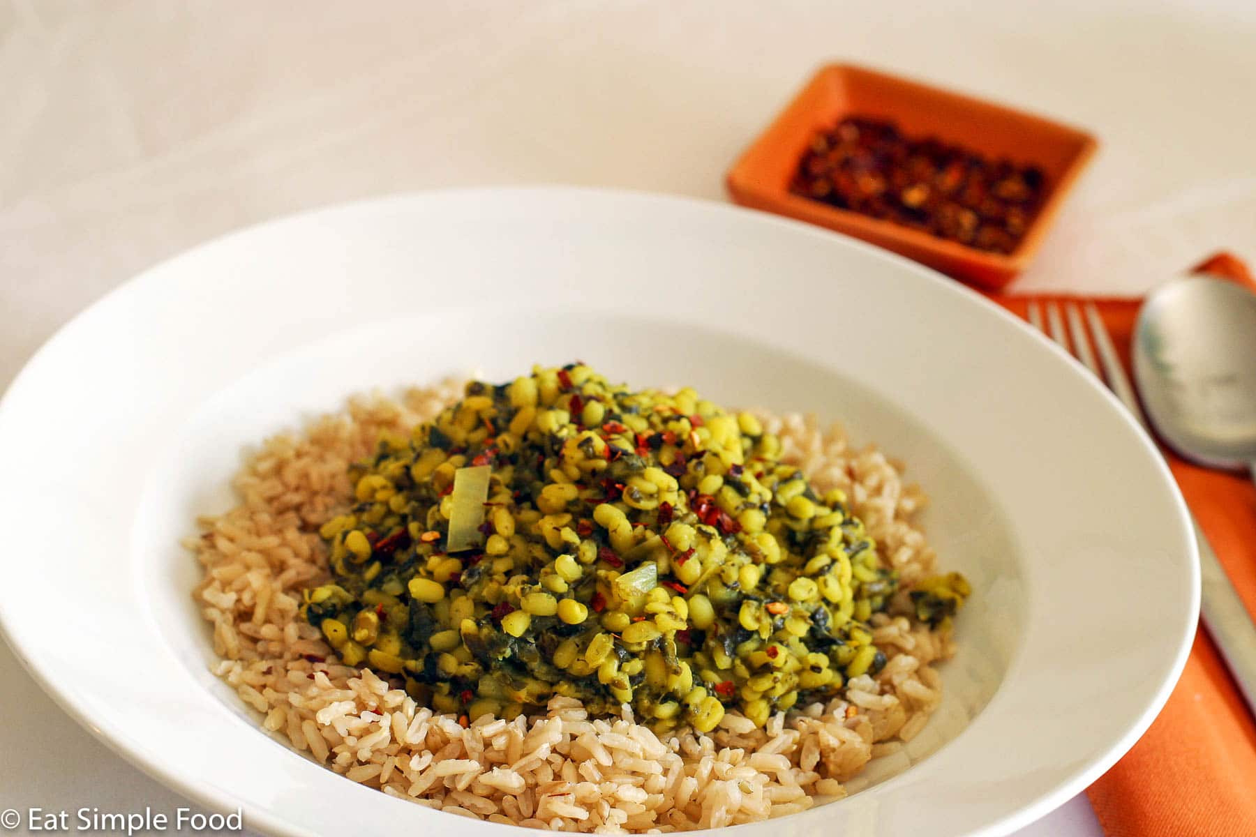 Mung Beans piled on top of brown rice on a shallow white bowl with red pepper flakes on the side.
