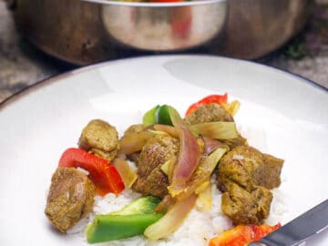 White plate of browned chunks of lamb with seared red onions and red and green peppers. Pot behind contains same ingredients.