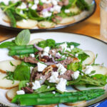 2 white plates with a layer of sliced cooked potatoes, blanched green asparagus spears, spinach, and seared lamb with crumbled feta garnish. Close up.