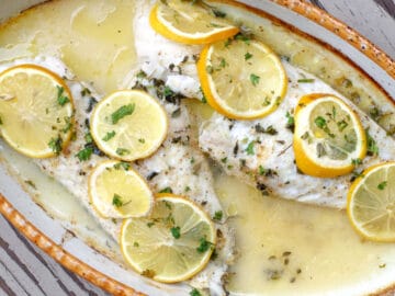 Top view. 2 fillets of white fish in a lemon butter sauce with thin sliced lemons over the top of the fish and a chopped parsley garnish.