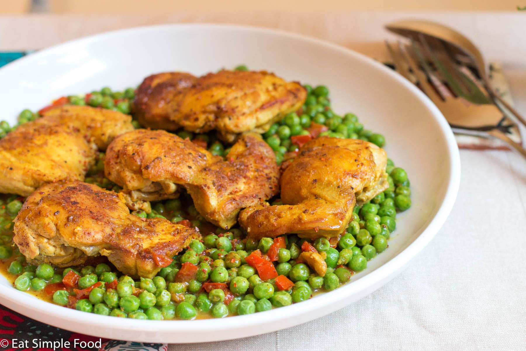 Top view. White deep platter with 5 browned cooked chicken thighs on a bed of bright green peas and cooked peppers. Serving silverware on side.