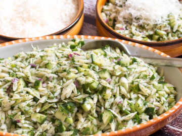 2 bowls of orzo pasta in a green herb sauce with diced cucumbers, red onions, and green onions. One bowl has shredded parmesan and a spoon. Side view.
