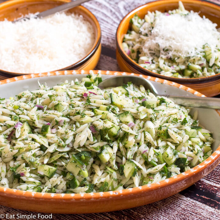 2 bowls of orzo pasta in a green herb sauce with diced cucumbers, red onions, and green onions. One bowl has shredded parmesan and a spoon. Side view.