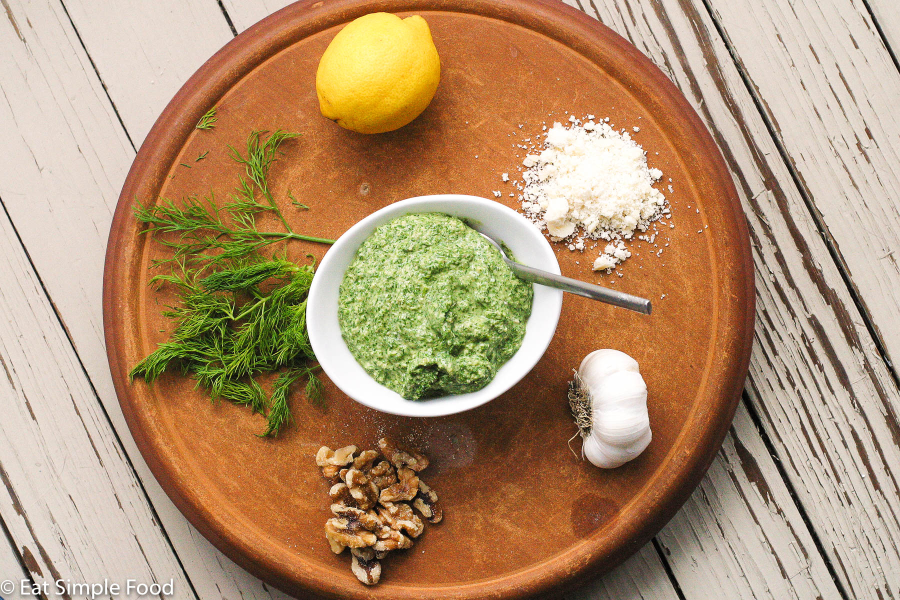 White bowl of green dill pesto with a spoon sticking out in the middle of a cutting board with a lemon, pile of grated Parmesan cheese, garlic bulb, handful of walnuts and fresh dill sprigs.
