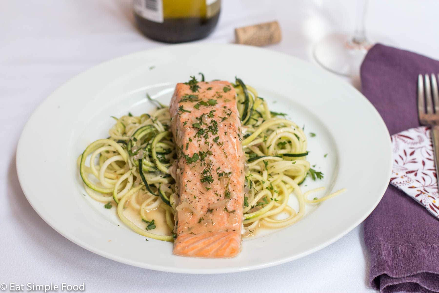 Salmon filet on a white plate on top of zucchini noodles with parsley and caper garnish. wine in back drop with white table cloth.