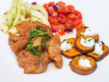 Sliced Pork Tenderloin browned with cilantro garnish on a white plate with roasted sweet potato coins with sour cream and chives, tomato salsa, and cucumber salad