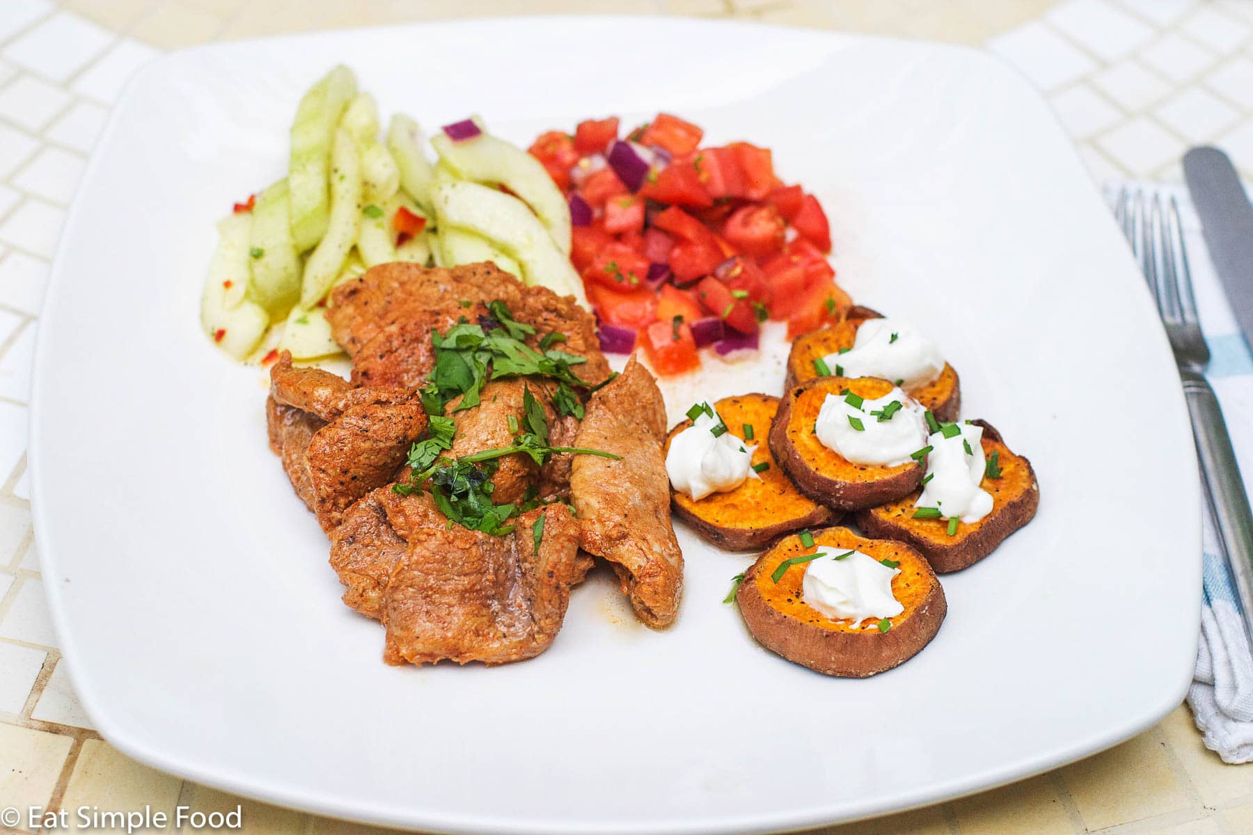 Sliced Pork Tenderloin browned with cilantro garnish on a white plate with roasted sweet potato coins with sour cream and chives, tomato salsa, and cucumber salad