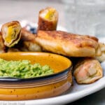 Deep Fried Egg Rolls filled with green chiles, cheese, and mangos stacked on a white platter and served with a bowl of guacamole.