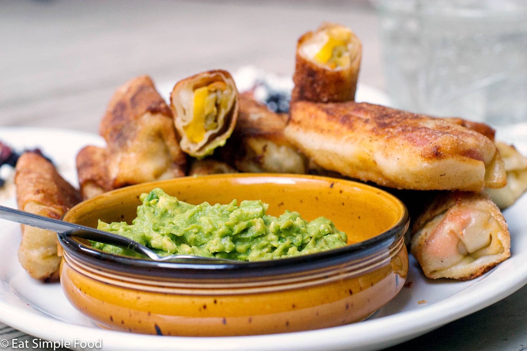 Deep Fried Egg Rolls filled with green chiles, cheese, and mangos stacked on a white platter and served with a bowl of guacamole.