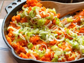 Mashed Carrots and Sliced Leeks in a cast iron pan with a wood spoon on a brown counter. side view