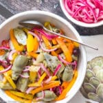 Top view of artichoke salad with kalamata olives, sliced orange peppers, capers and a bowl of pickled red onions on the side.