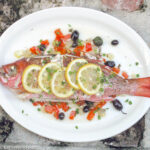 Cooked Red Snapper with red peppers and sliced olives on a white plate with sliced lemon wedges on top and a parsley garnish.