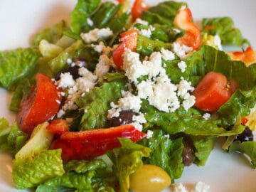 Romaine lettuce chunked, chunks of tomatoes, red peppers, whole olives and crumbled feta cheese. On a white plate.