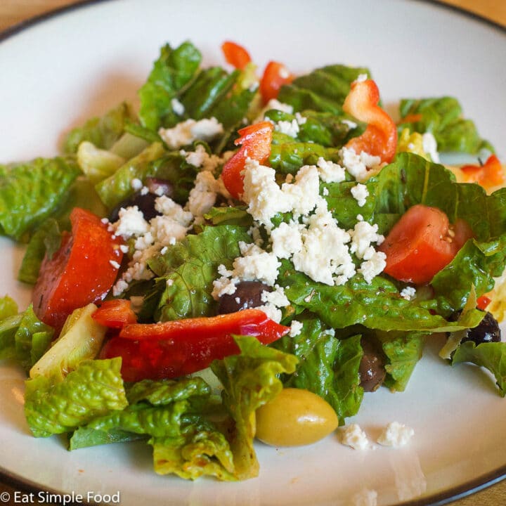 Romaine lettuce chunked, chunks of tomatoes, red peppers, whole olives and crumbled feta cheese. On a white plate.