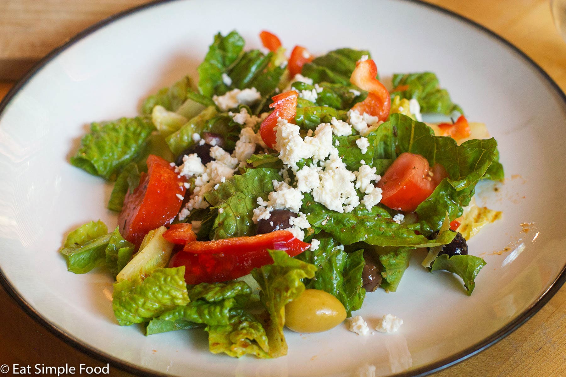 Romaine lettuce chunked, chunks of tomatoes, red peppers, whole olives and crumbled feta cheese. On a white plate.