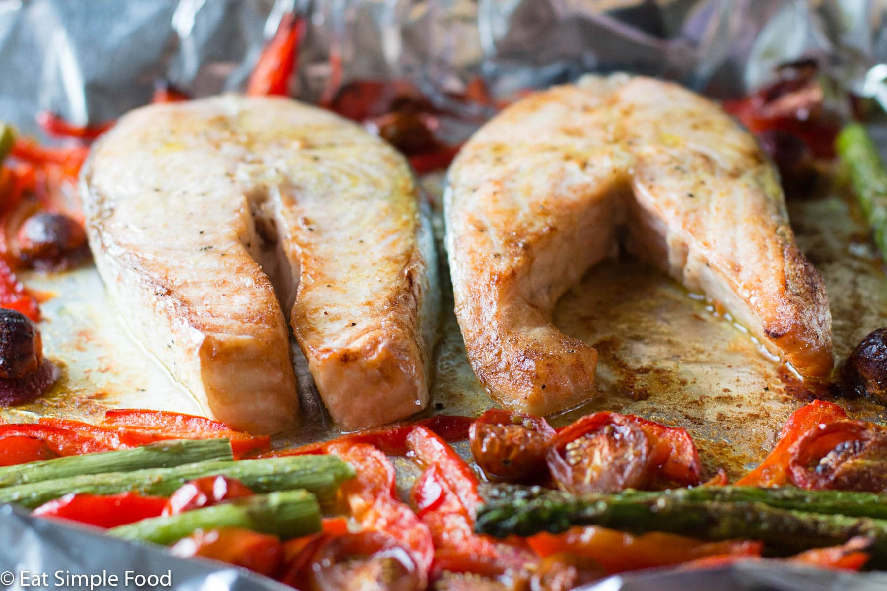 2 pieces of raw salmon fillets on an aluminum sheet pan with asparagus, sliced red peppers, and halved cherry tomatoes.