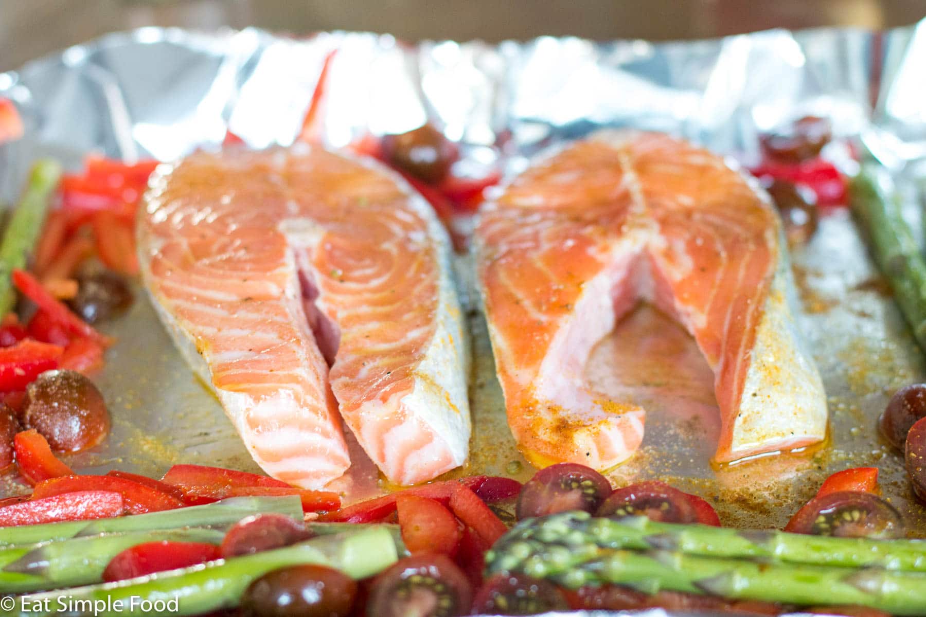 2 pieces of raw salmon fillets on an aluminum sheet pan with asparagus, sliced red peppers, and halved cherry tomatoes.