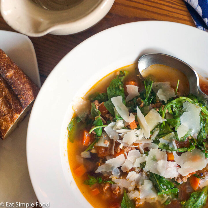 White bowl of tomato based soup with sausage, carrots,arugula, white beans, and garnished with thin slices of Parmesan cheese. Spoon and baguette on the side.