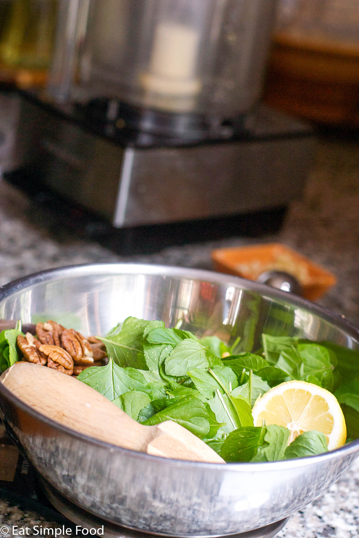 Stainless steel bowl of raw ingredients for mint pesto: mint, lemon, pecans, garlic. Wood handheld lemon juicer in bowl.