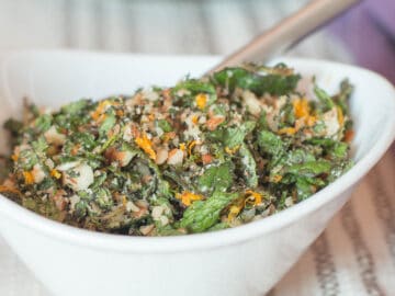 White oval bowl with orange zest, sliced mint, minced garlic and chopped almonds with a spoon hanging out on a tan and white striped table cloth.