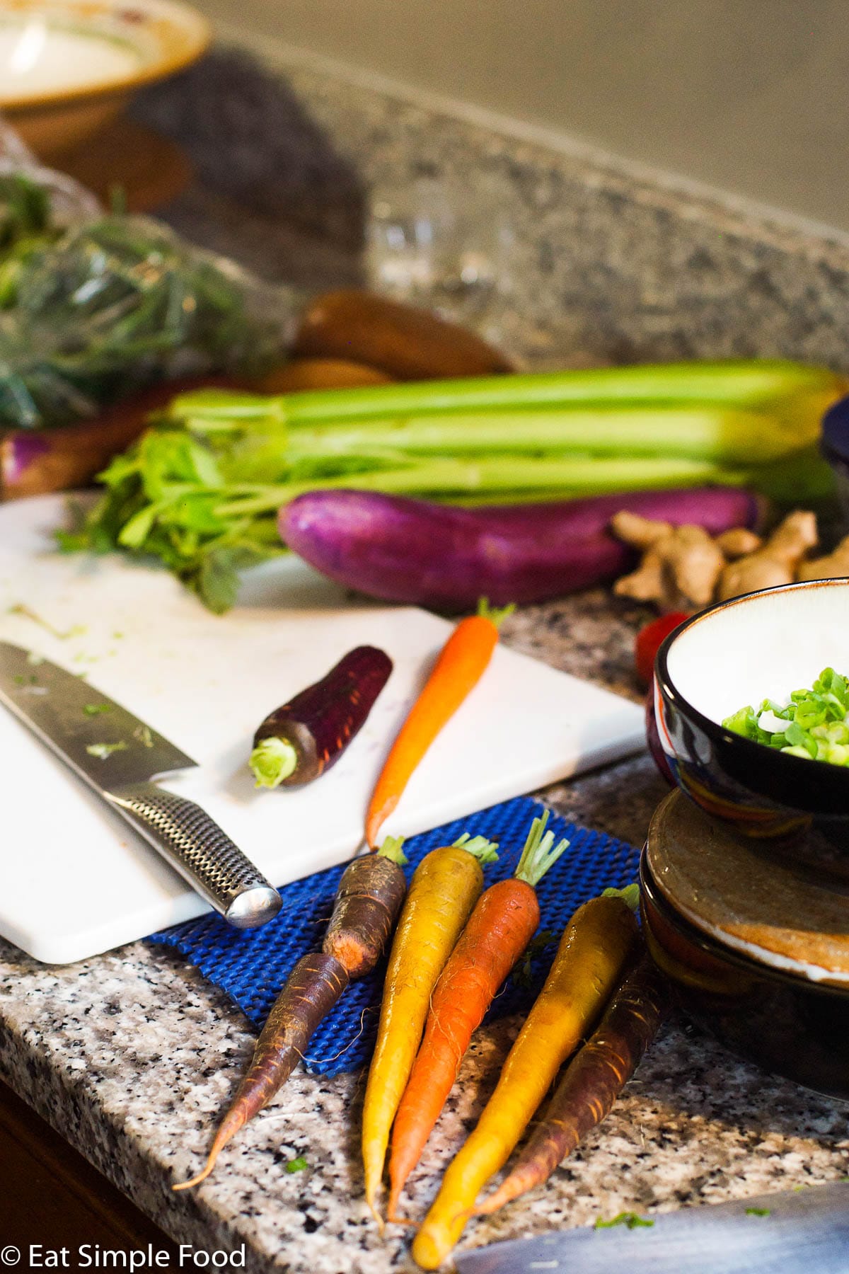 Colorful carrots, celery, and eggplant near a white cutting board with a chef's knife getting ready to be cut.