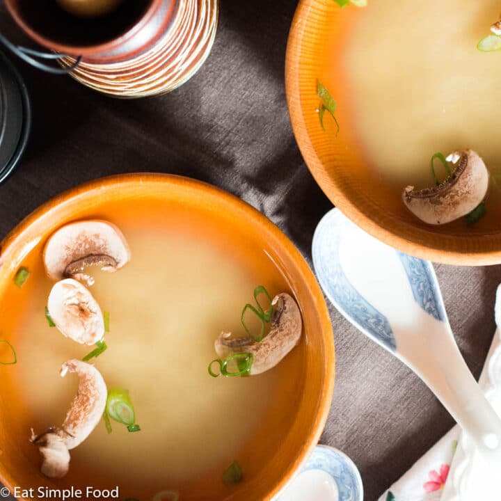 2 small wood bowls filled with brown miso soup with green onions and sliced mushrooms floating. 2 traditional Japanese spoons. Top view.
