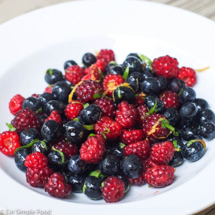 White shallow bowl filled with raspberries, blueberries, and blackberries. Garnished with orange zest and sliced mint.