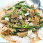 White shallow bowl filled with white rice and topped with potato, eggplant, green bean tan curry with a sliced green onion garnish.