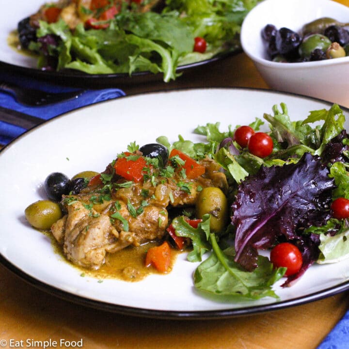2 plates of browned chicken with brown sauce, chunked red peppers, whole green and black olives and a mixed green and purple salad with whole cherry tomatoes.