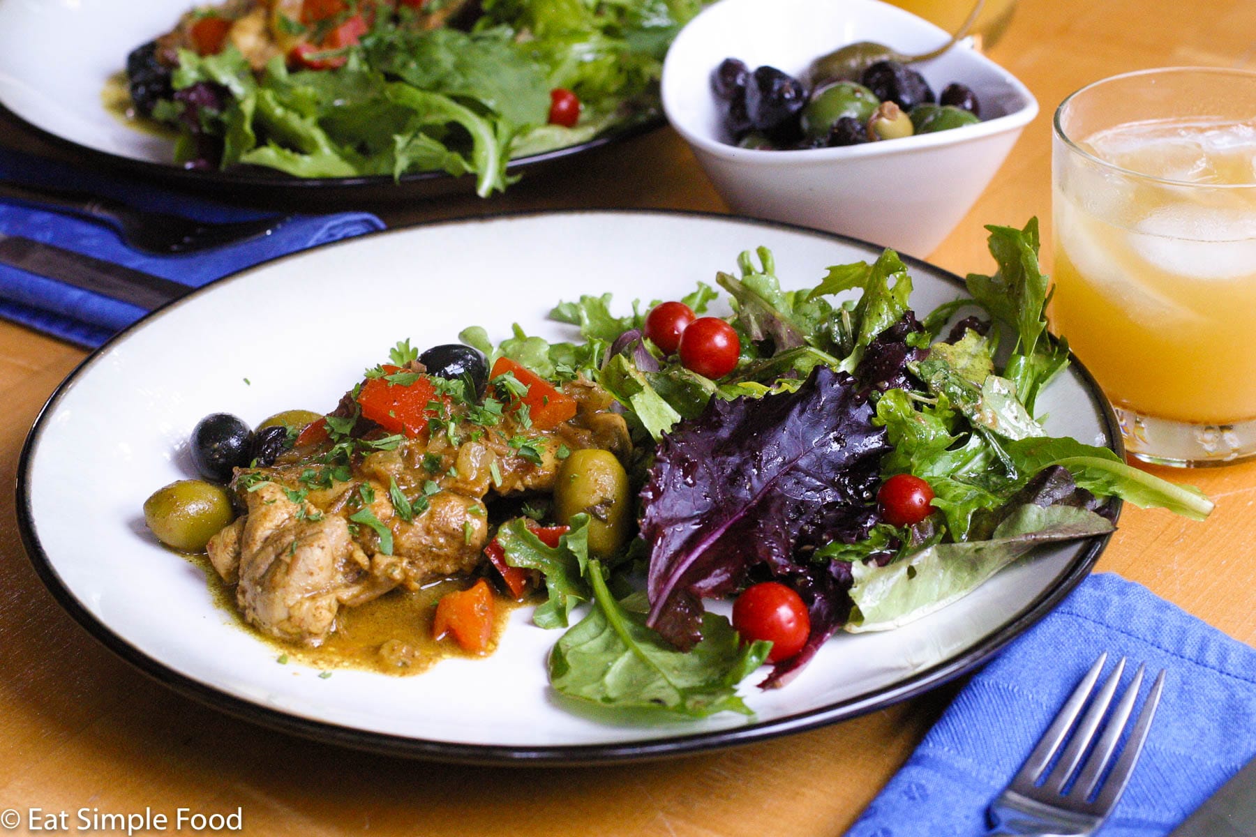 2 plates of browned chicken with brown sauce, chunked red peppers, whole green and black olives and a mixed green and purple salad with whole cherry tomatoes.