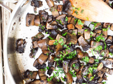 Stainless steel pan full of cooked Portobello Mushrooms with parsley garnish and a wood spoon sticking out of it.
