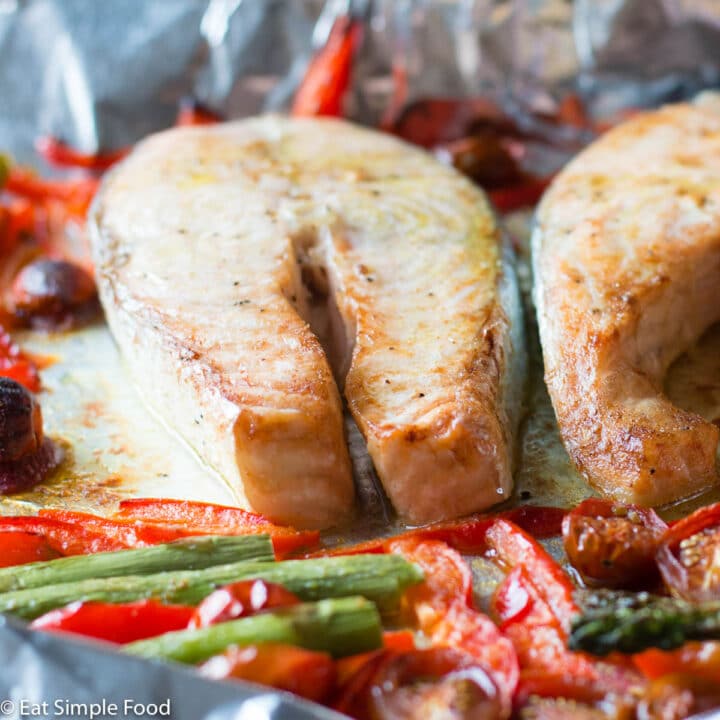 2 pieces of raw salmon fillets on an aluminum sheet pan with asparagus, sliced red peppers, and halved cherry tomatoes.