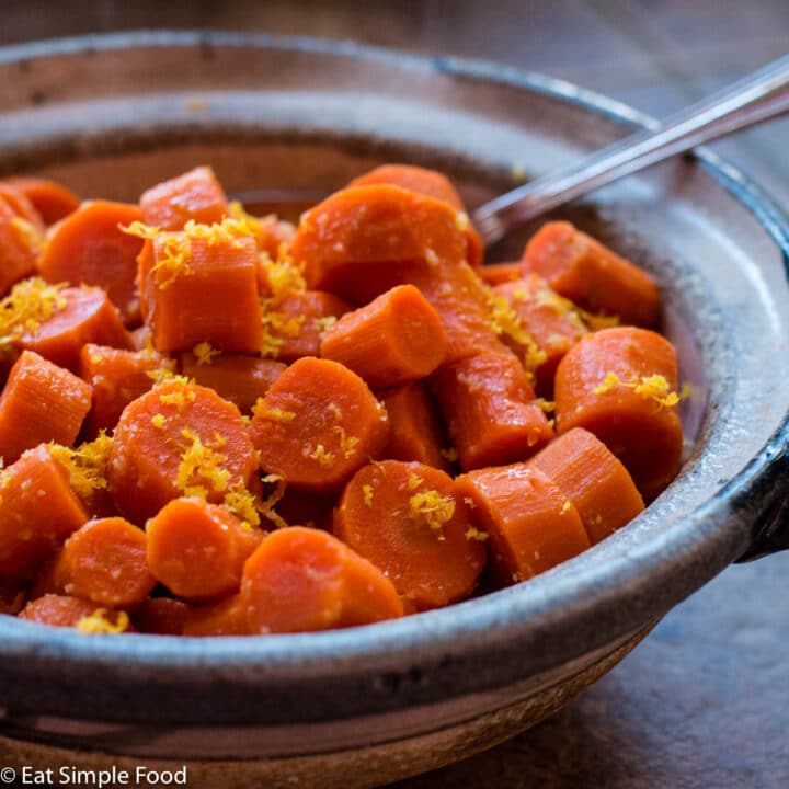 Side view of chunked carrots in a tan bowl with orange zest garnish and a spoon out the side..
