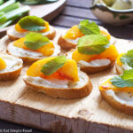 Crostini (little toasts) topped with spreaded mascarpone, a peach slice, and a mint leaf. Several crostini's ona wood cutting board.