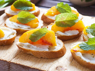 Crostini (little toasts) topped with spreaded mascarpone, a peach slice, and a mint leaf. Several crostini's ona wood cutting board.