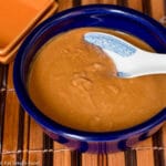 Blue Bowl of Brown Peanut Sauce with a white chinese style spoon in it. On a bamboo placemat.