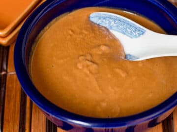 Blue Bowl of Brown Peanut Sauce with a white chinese style spoon in it. On a bamboo placemat.