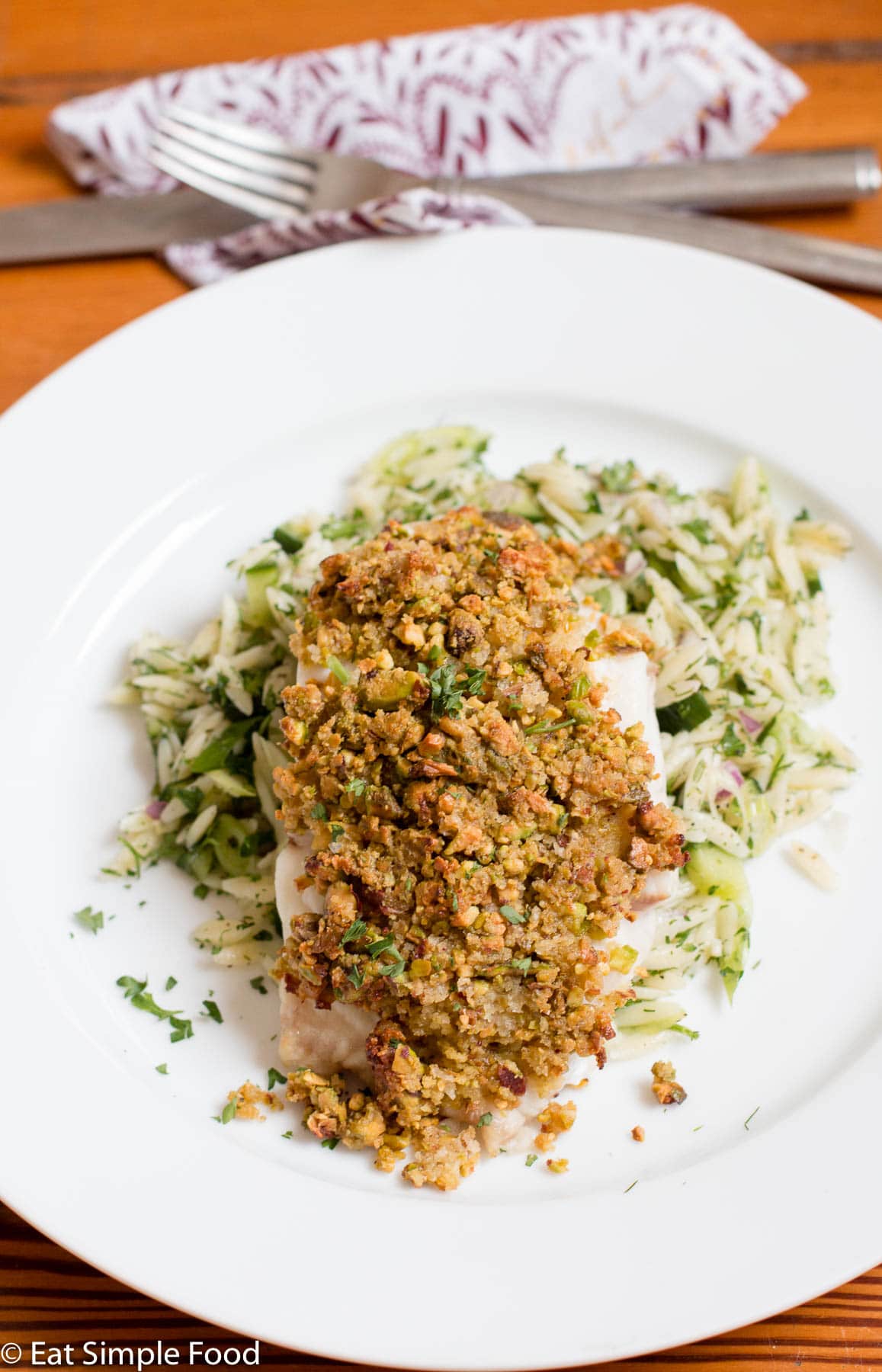 A cooked chicken Breast coated in browned processed pecans. Garnished with parsley and on a bed of orzo with herbs on a white plate.