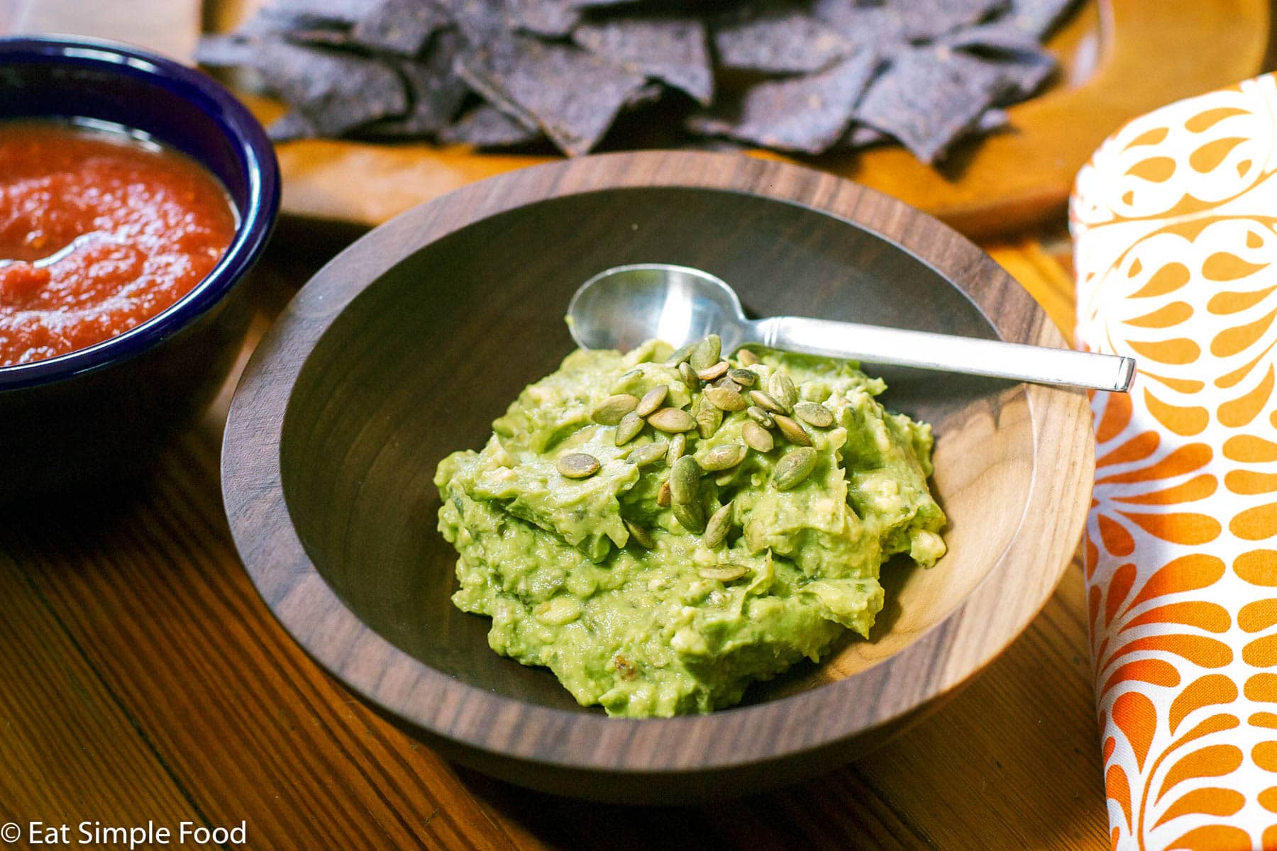 Guacamole in a wood bow with toasted pepita garnish with a spoon in bowl. Blue tortilla chips and bowl of salsa in background.