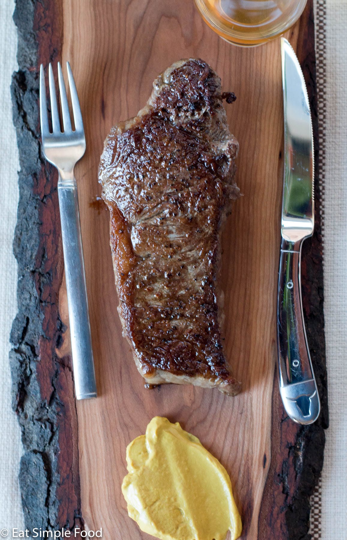 New York Strip Steak golden brown on a wood cutting board with a lived edge. fork on the left. steak knife on the right. sharp yellow mustard beneath.