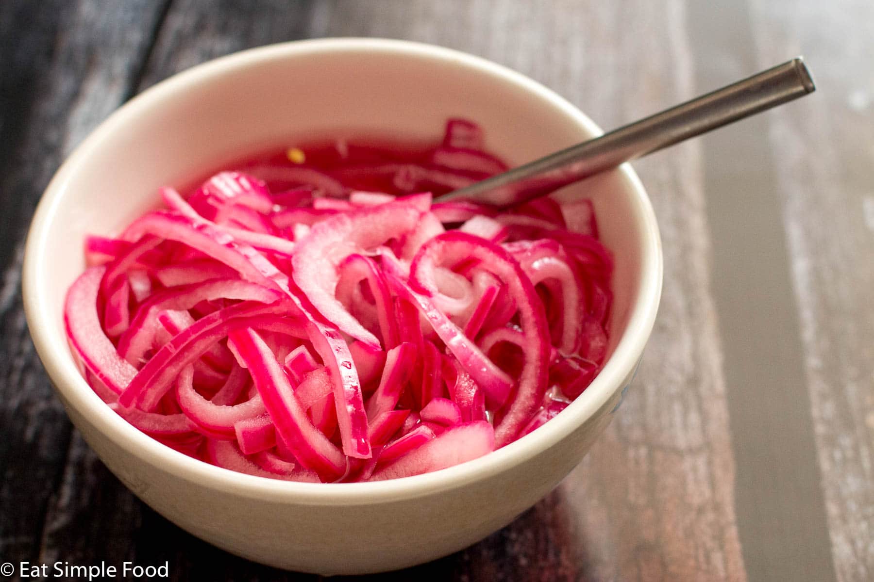 White bowl of sliced thin and pickled red onions (pink hue). with a silver spoon out of the bowl on a dark wood table.