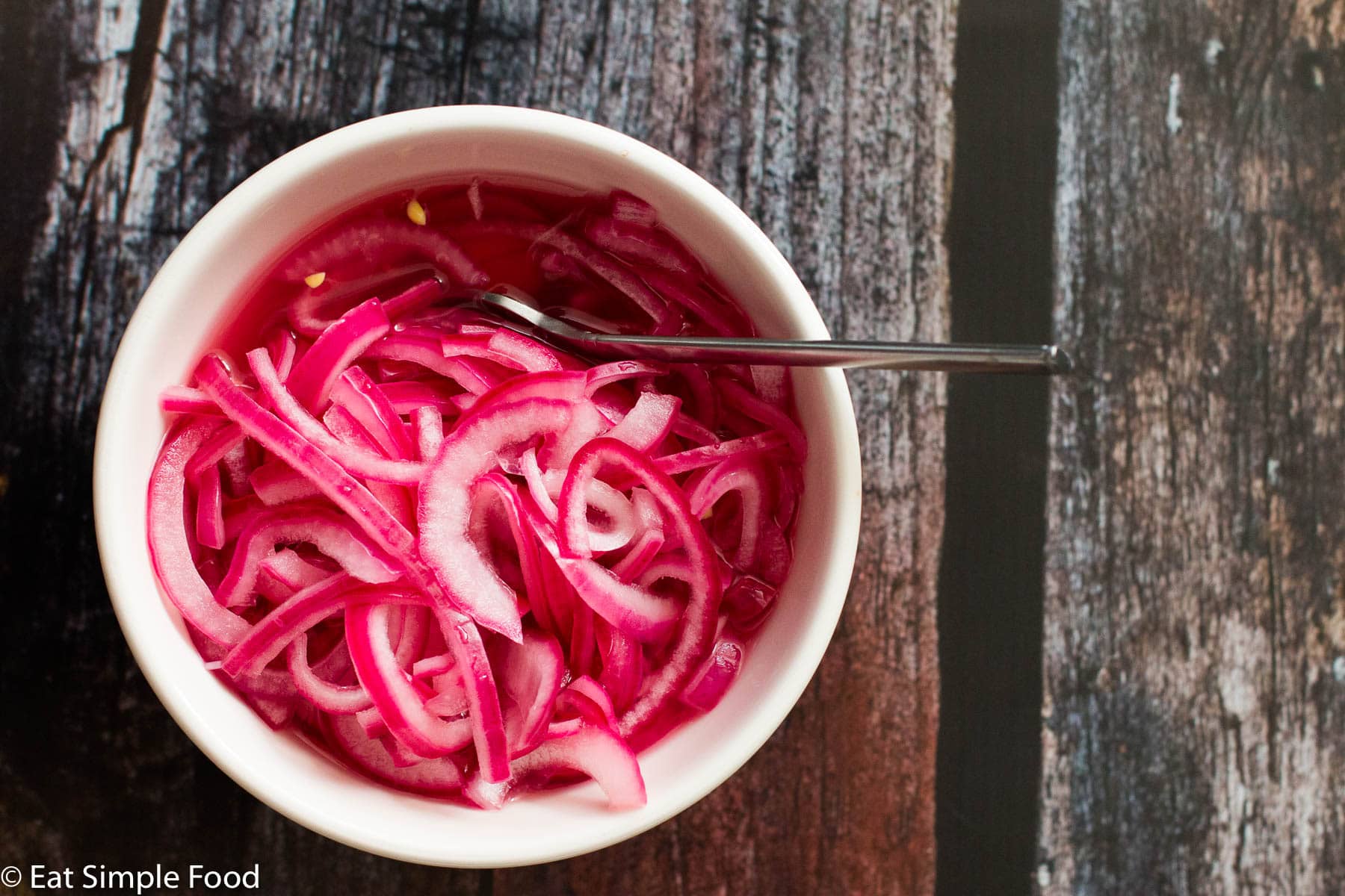 White bowl of sliced thin and pickled red onions (pink hue). with a silver spoon out of the bowl on a dark wood table. Top view.