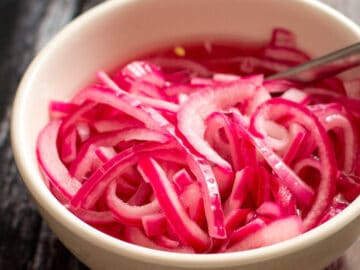 White bowl of sliced thin and pickled red onions (pink hue). with a silver spoon out of the bowl on a dark wood table.