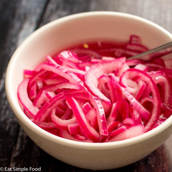 White bowl of sliced thin and pickled red onions (pink hue). with a silver spoon out of the bowl on a dark wood table.